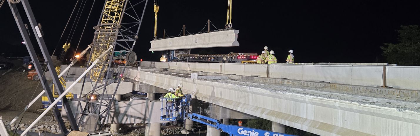 Nighttime photo of bridge work along a four-lane divided highway. Construction workers are visible along with a large crane holding a concrete girder.