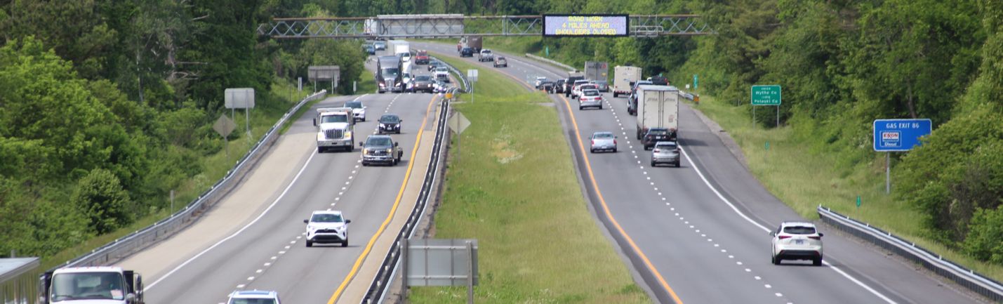 Four-lane highway with a center grass median and a digital message sign in the background. A mix of cars and trucks are on the travel lanes, moving directly toward or away from the camera.
