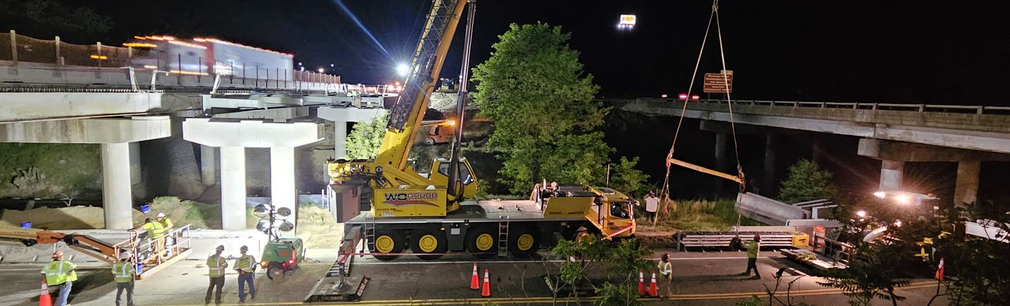 Nighttime photo of construction crews using a large crane to place a steel beam on new bridge piers. Traffic is visible to the left side of the image.