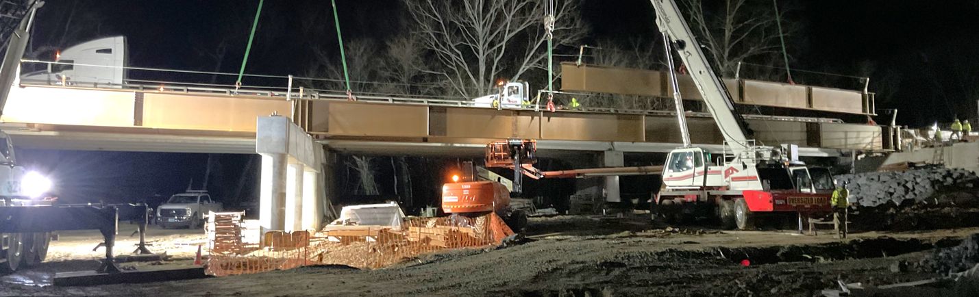 Nighttime photo of heavy construction equipment placing steel beams for a new highway bridge