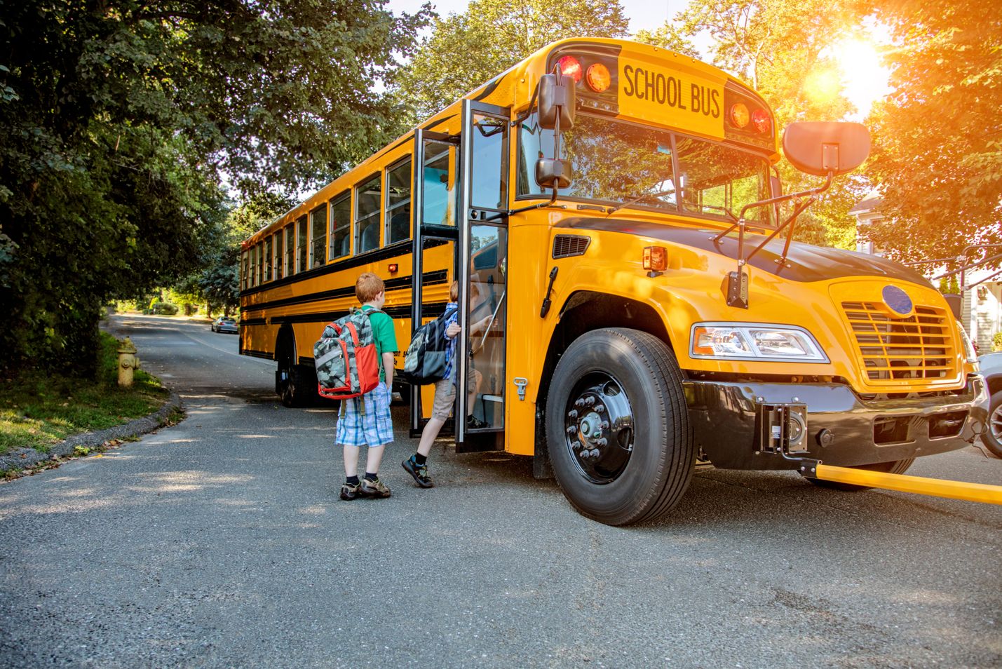 Two children board a yellow school bus stopped on the road with its gate extended