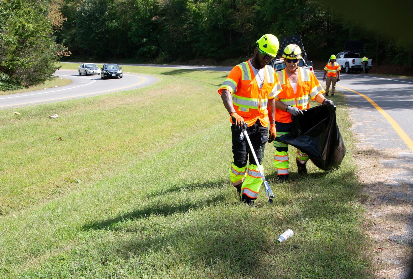 Two people dressed in bright helmets and clothing pick up litter along the side of the road.