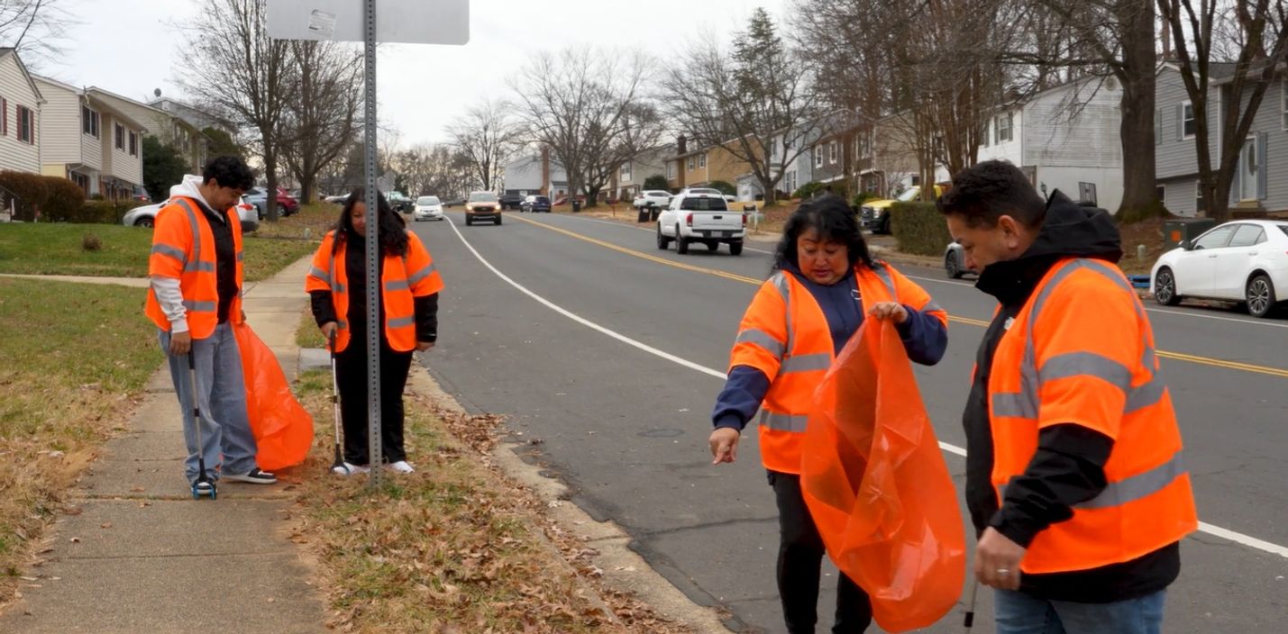 Four people wearing reflective vests pick up litter along a roadside.