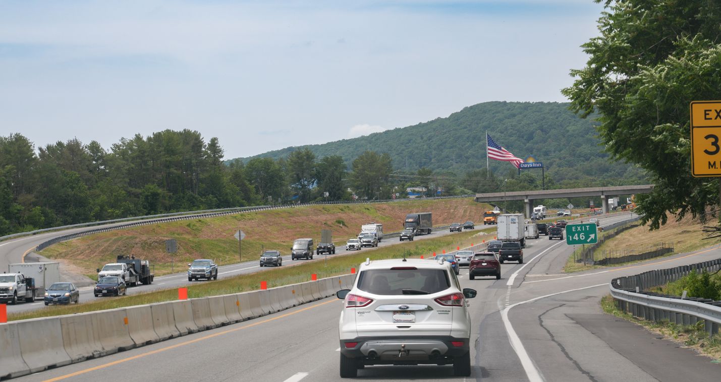 Vehicles travel in four lanes of highway traffic with an American flag in ahead with mountains behind it.