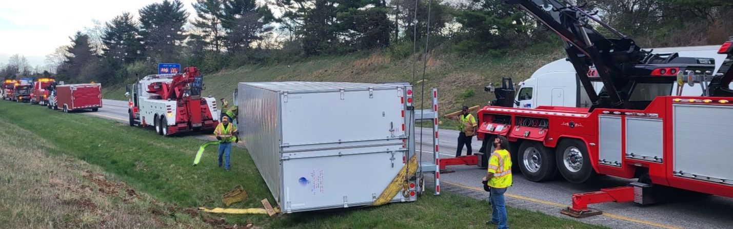 Photo of an overturned trailer along a multi-lane highway in a rural area. A crane, heavy-duty wreckers and other equipment and personnel are working on the trailer. Cloudy skies and some trees are in the background.