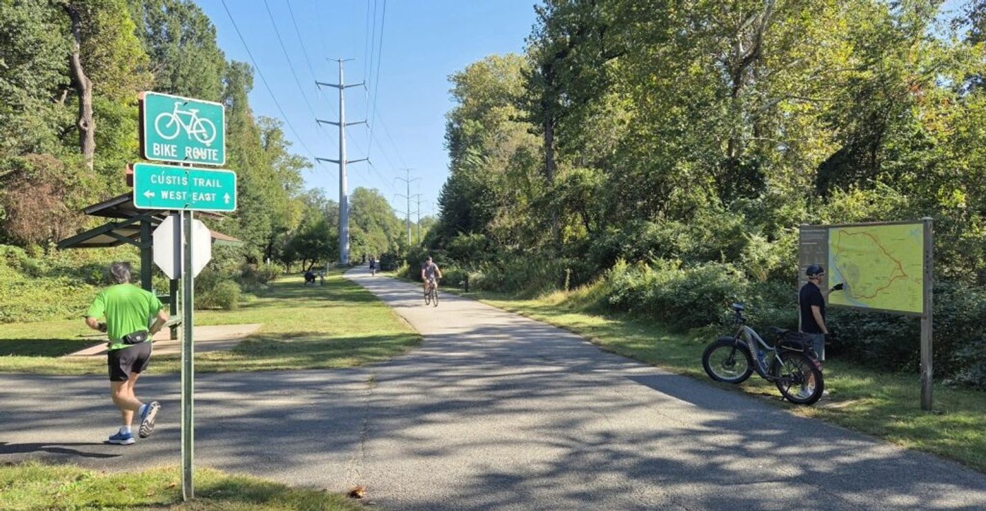 At an intersection of a multiuse bike trail, a man is jogging to the left, a dismounted cyclist is on the right looking at a map, and another cyclist is in the center if the image riding