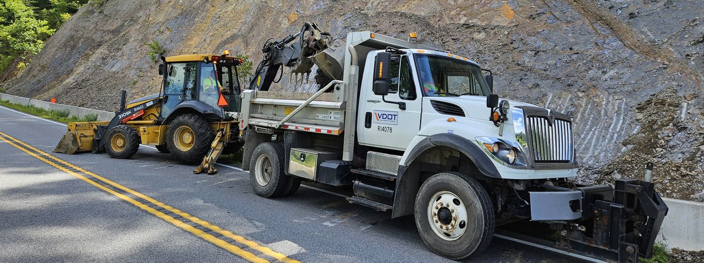 A backhoe and dump truck moving loose rock from the bottom of a steep slope. The heavy equipment is blocking one lane of a roadway.