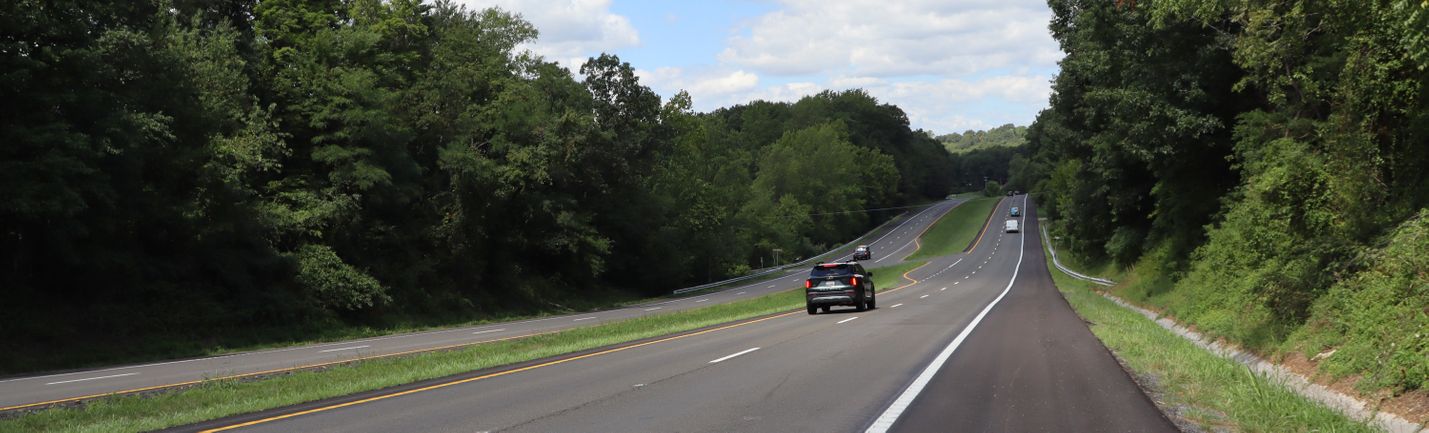 A four-lane highway with light traffic on a rural area. Right side of roadway shows fresh pavement and lane-marking paint. Partly cloudy skies and leaf-covered trees are in the background.