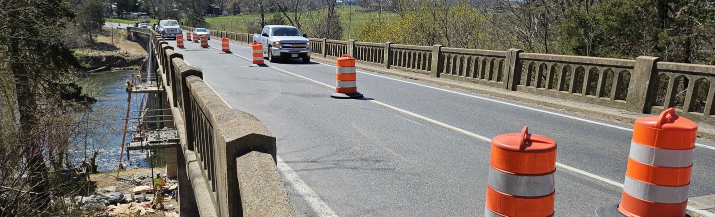Vehicles moving along a bridge under construction over a river. Orange barrels restrict traffic to a single lane.