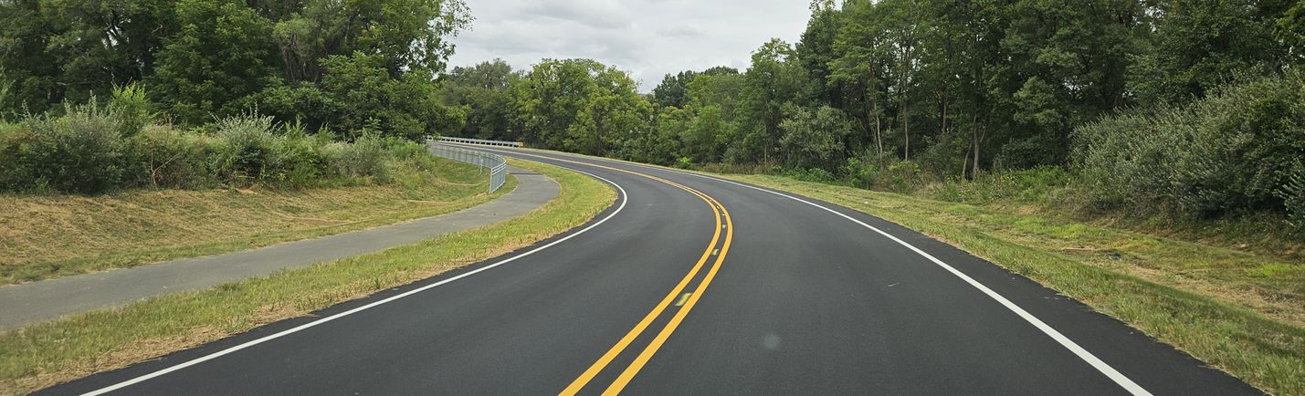 In-vehicle photo of a two-lane roadway curving off to the left. A shared-use path runs parallel to the road on the left. Also visible are trees and cloudy skies.