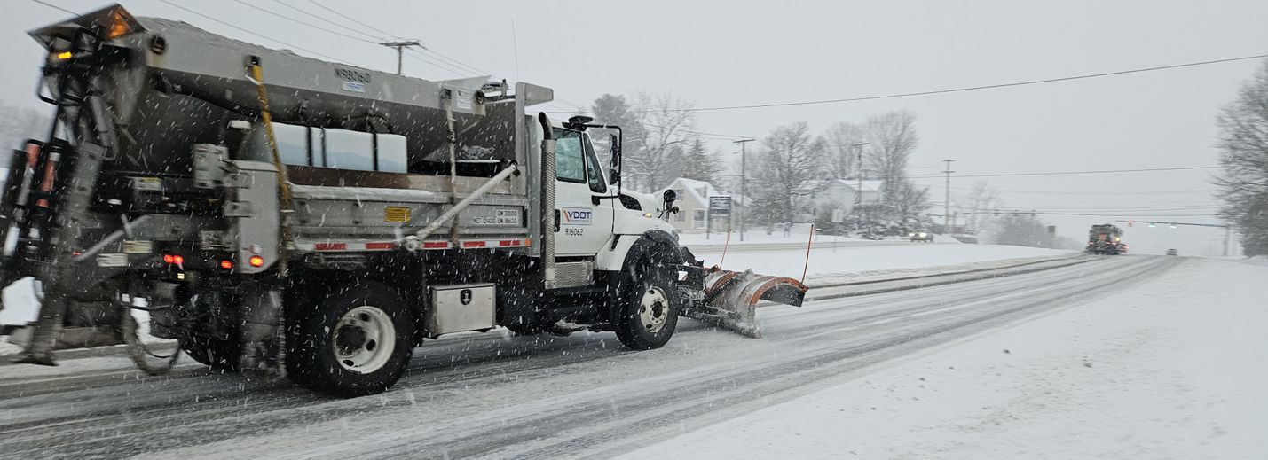 VDOT truck plowing snow on a four-lane roadway, with snow continuing to fall from an overcast sky