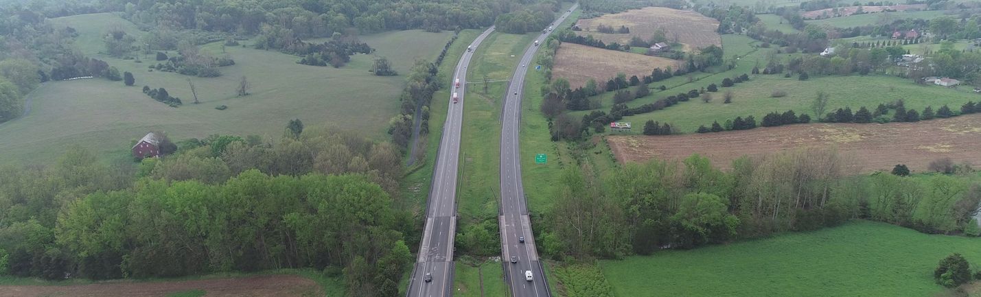 Aerial view of interstate highway in a rural area, showing bridges over a stream and an uphill grade. Trees, a few buildings and overcast skies are in the background.