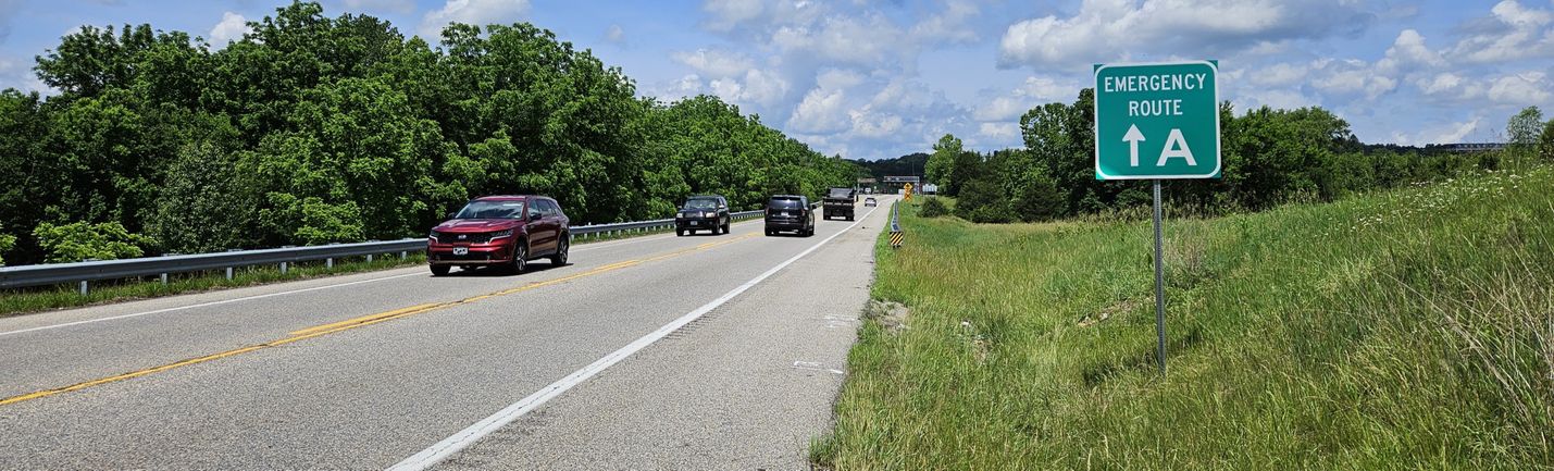 Moderate traffic on a two-lane rural road on a sunny summer day. At the right is a green square sign with white lettering that reads, Emergency Route A