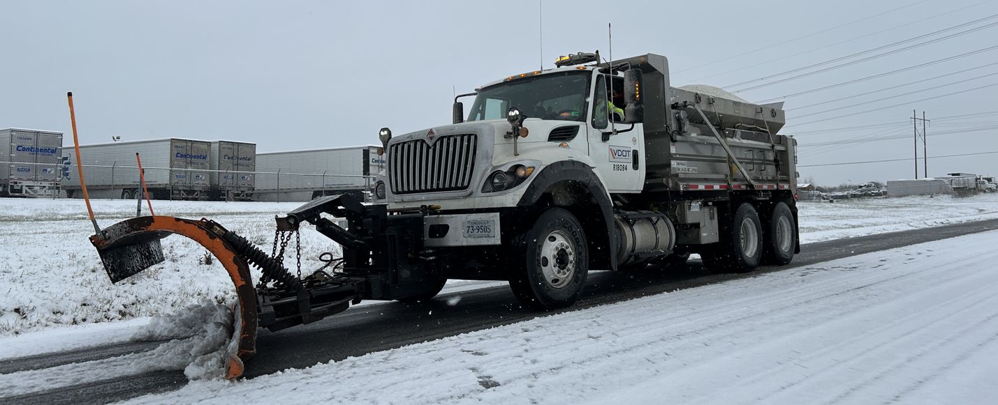 Snowy-day photo of a plow clearing a two-lane road with parked tractor trailers in the background.