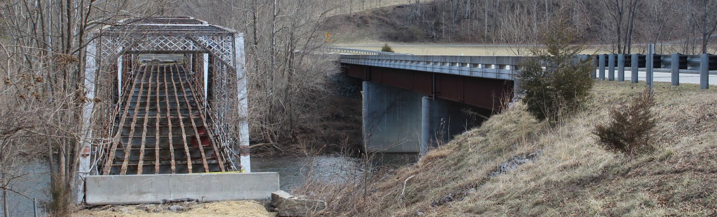 Old truss bridge next to a more modern concrete bridge over a river. Background shows bare trees and overcast skies.