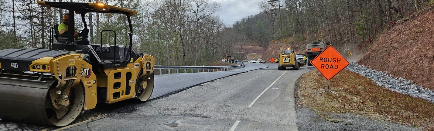 Work zone on a two-lane, curving roadway with heavy equipment including a steamroller. Orange signs, trees and an overcast sky are in the background.