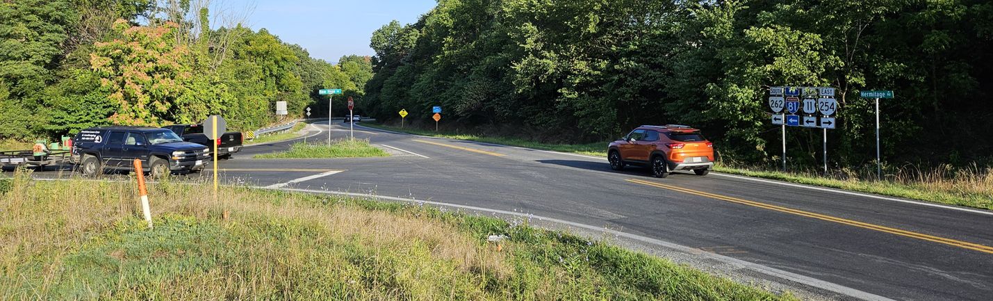 Wide-format photo showing the rural intersection of a pair of two-lane roads, with moderate traffic and green trees in the background on a sunny day.