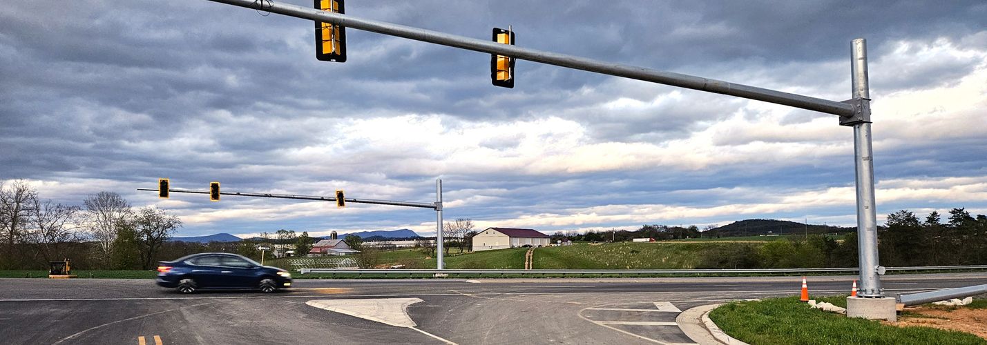 Photo of a rural intersection with a partial traffic signal above. The signals boxes are covered in black. The background shows a cloudy sky and green fields.