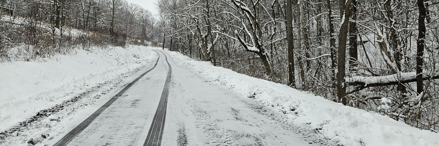 A wide-format photo of a rural road with a thin layer of snow cover, and vehicle tracks running down the center. Snow-covered ground and trees are in the background.