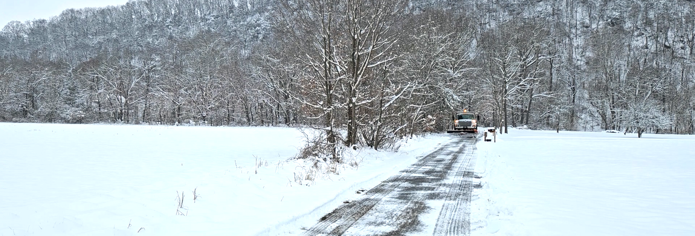 A snowplow approaching from the distance along a rural road partly covered with snow. Mountains and trees, all snow-covered, are visible