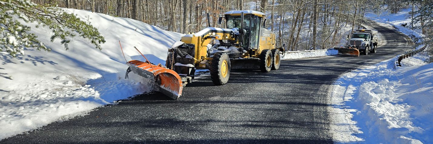 VDOT motor grader and snowplow pushing snow and ice from the edge of a secondary road. Bare trees and a clear blue sky are visible.