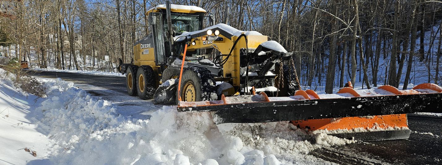 Motor grader plowing snow along the side of a secondary road. Trees and a clear blue sky are visible in the background.