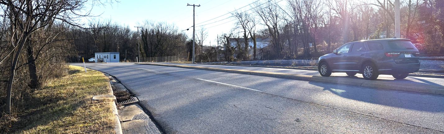 Wintertime photo of a four-lane roadway with a single vehicle moving right to left. Bare trees and a few buildings are on the sides of the road.
