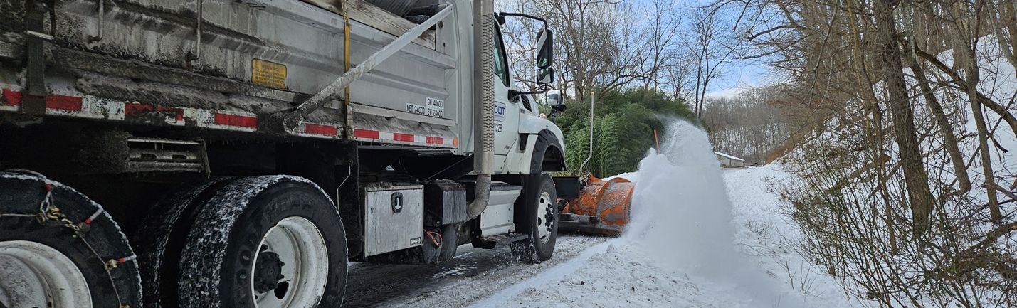 Photo of a VDOT snowplow pushing snow along a back road in a wooded area under mostly sunny skies. The truck is moving away from the camera, and a rooster tail of snow is visible off the end of the plow blade.