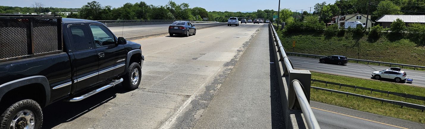 Photo of a multi-lane bridge over a four-lane divided highway on a sunny day with moderate traffic. Trees, a few buildings and blue skies are in the background.