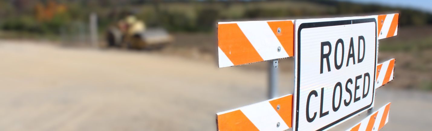 Stylized photo of a ROAD CLOSED barrier along a rural two-lane road.