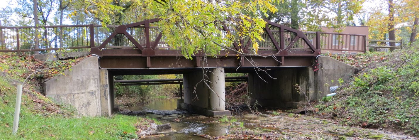 Side view of a small truss bridge carrying a roadway over a stream in a park-like setting