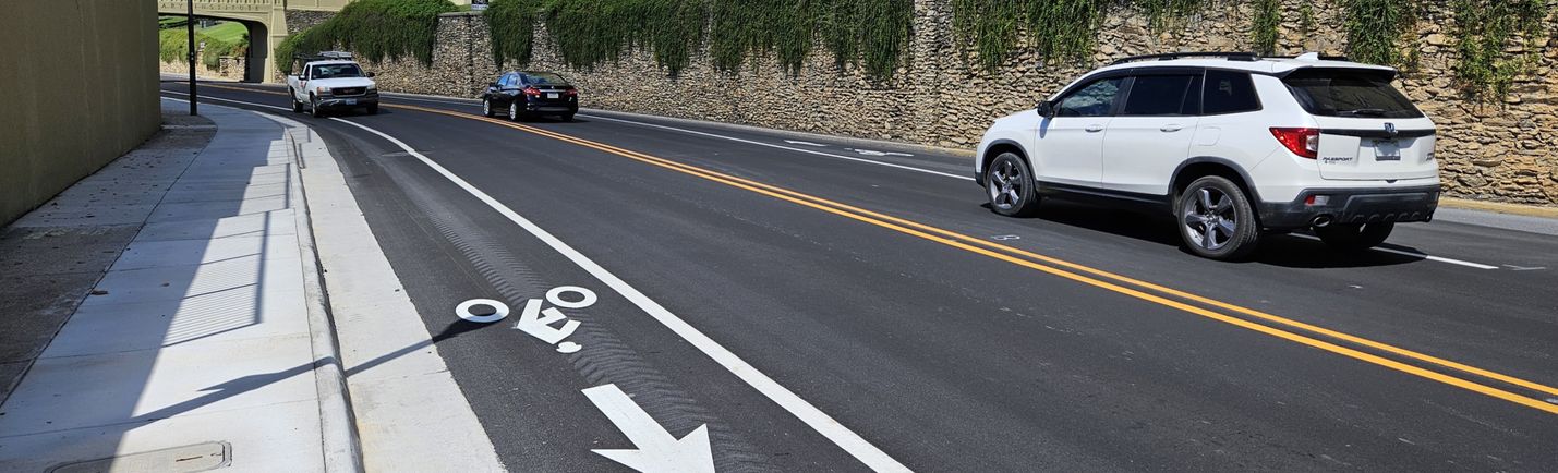 Sunny day photo of a two-lane roadway with moderate traffic. Buildings, a pedestrian bridge, sidewalks and bike lanes are in the shot.