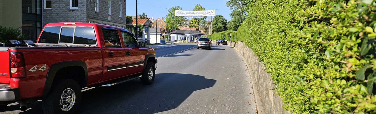 Traffic on a two-lane urban road with shrubbery along the right side of the roadway and blue skies overhead