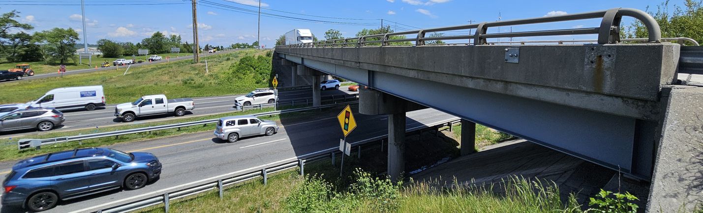 Photo of a tractor trailer driving on a bridge over a multi-lane highway with moderate traffic. Grass and other vegetation is in the foreground and background under a clear blue sky.