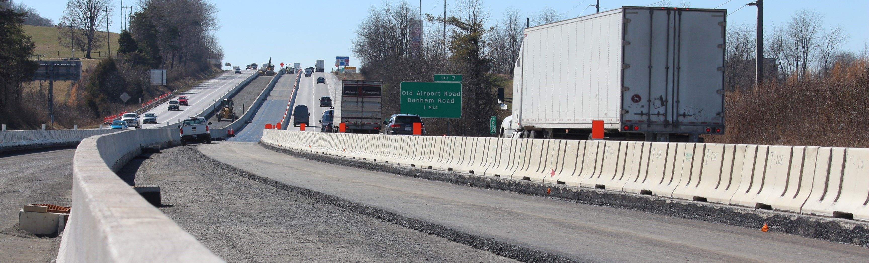 Interstate traffic moving through a work zone. Concrete barriers separate traffic from heavy equipment. Sections of new asphalt are visible.