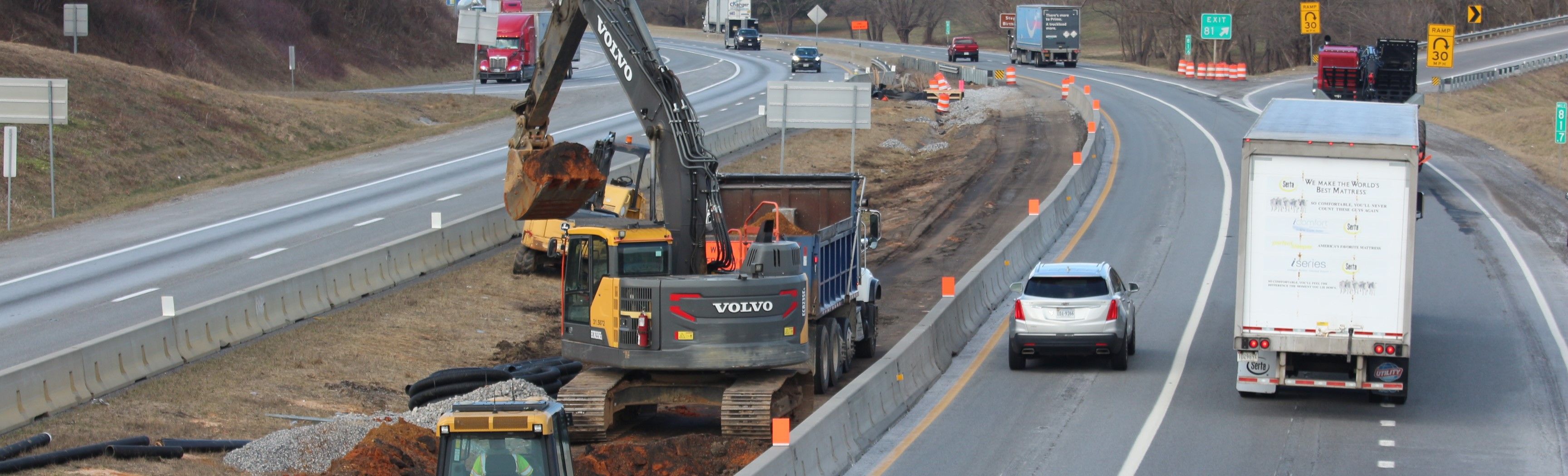 Excavation work taking place in the median between interstate travel lanes with traffic visible in both directions. At right is an off-ramp into a tight curve.