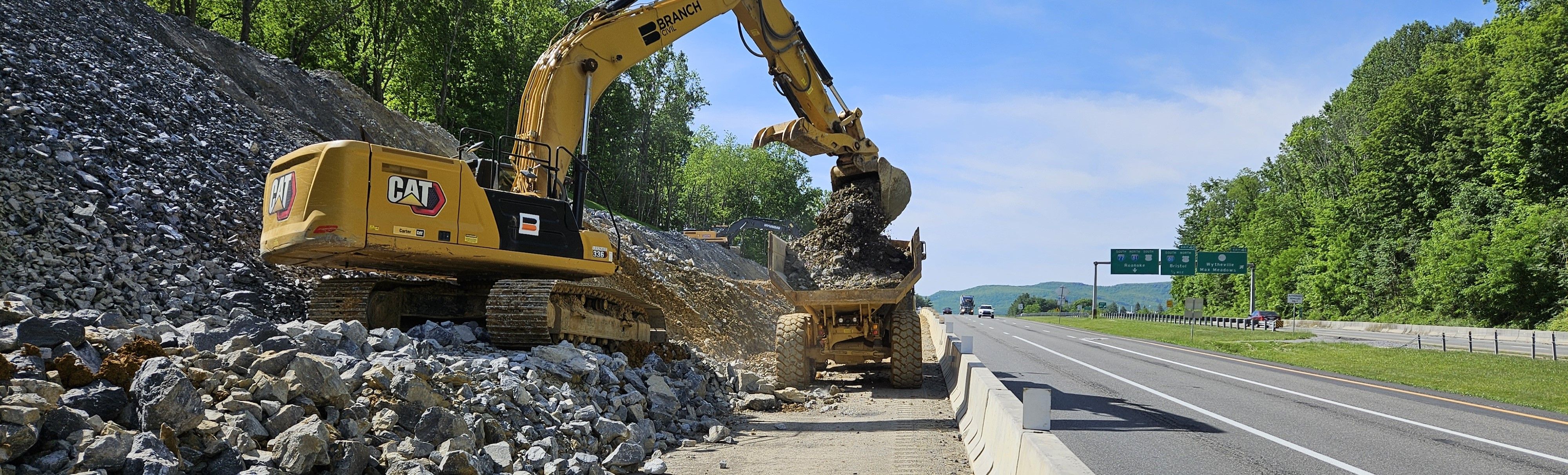 Sunny-day photo of a backhoe emptying a load of rock into a dump truck. The backhoe is on a steep embankment to the left. Concrete barrier separates the construction vehicles from a sparsely traveled highway.
