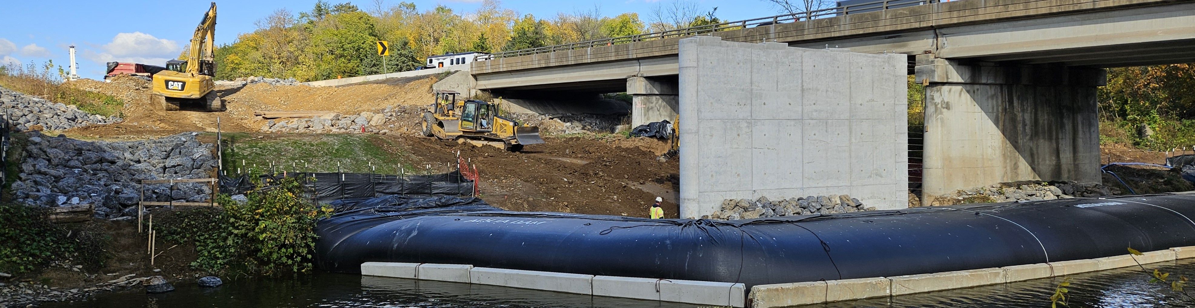 Photo of bridge construction over a waterway in the median of a multi-lane highway. Construction vehicles and equipment are visible along with traffic in the background and coffer dam in the foreground next to a concrete bridge pier.