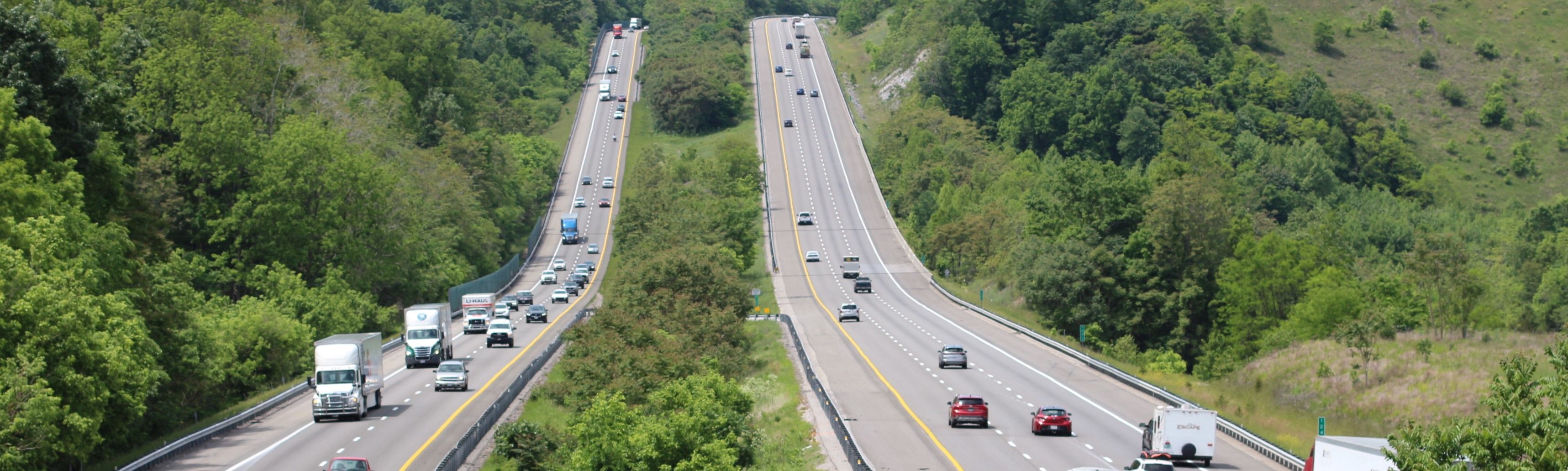Multi-lane highway in a rural area with a steep grade in the background. There is a moderate amount of traffic including personal vehicles and tractor trailers.