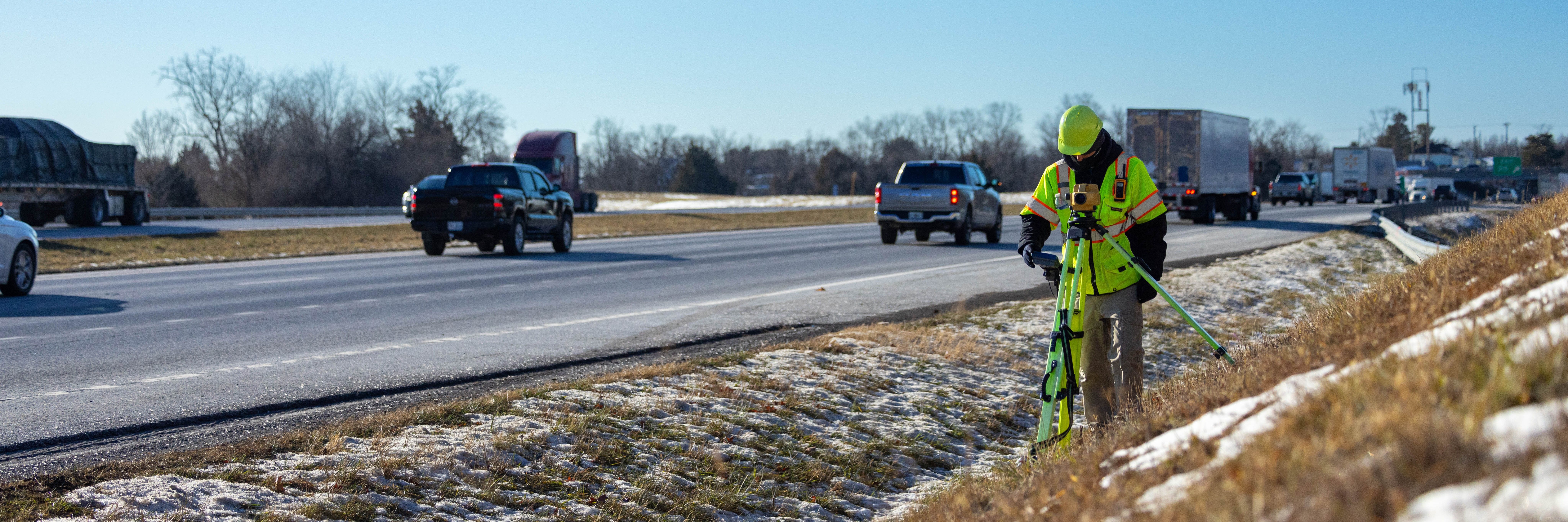 Surveyor in bright yellow clothing and a hard hat, working along interstate traffic. Photo captures a blue sky and patches of snow.