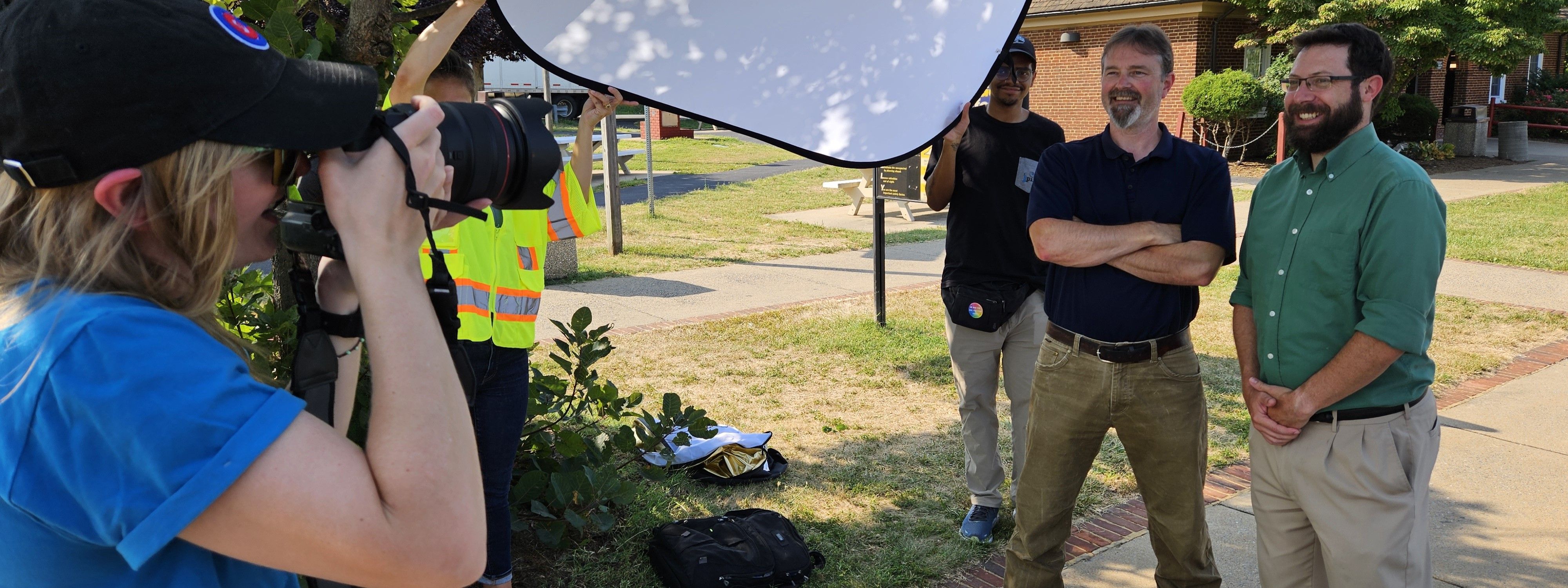 Two people standing in front of a third person pointing a long-lens camera at them. Also visible is a person holding a sunlight filter in the background.