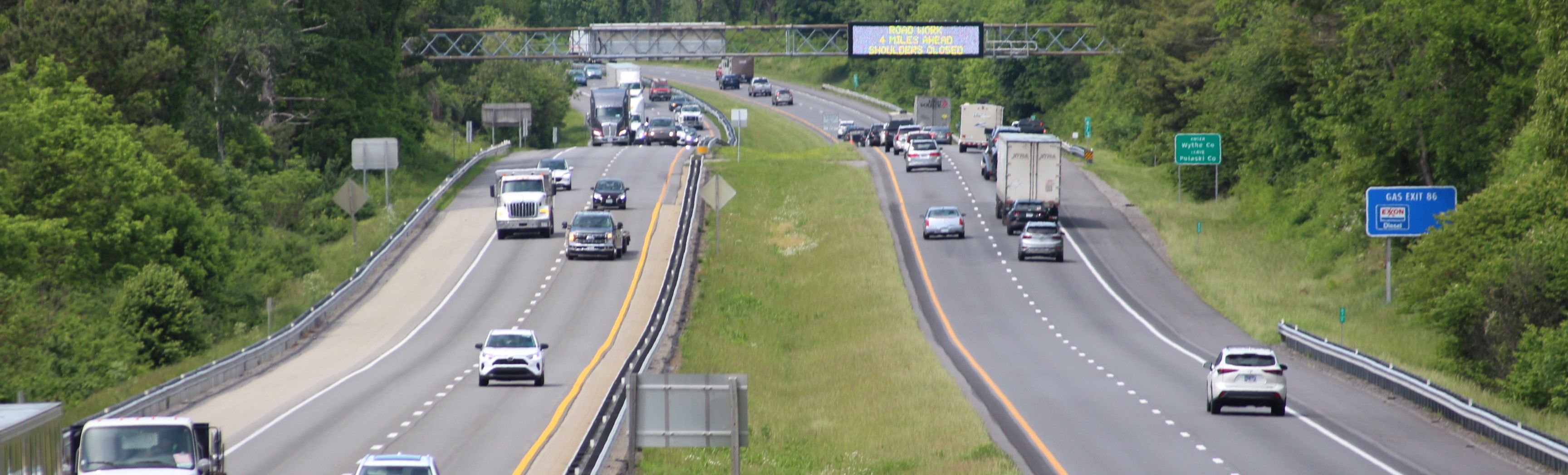 Four-lane highway with a center grass median and a digital message sign in the background. A mix of cars and trucks are on the travel lanes, moving directly toward or away from the camera.