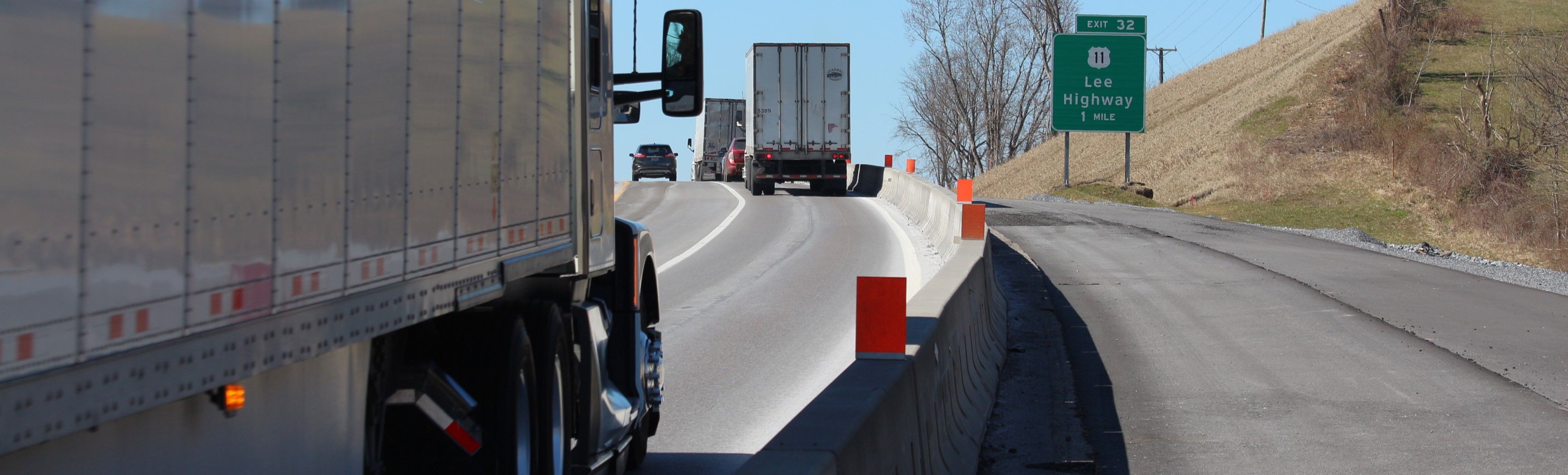 Trucks and other vehicles moving up a hill on an interstate highway. Concrete barriers separate traffic from a partially paved area to the right.