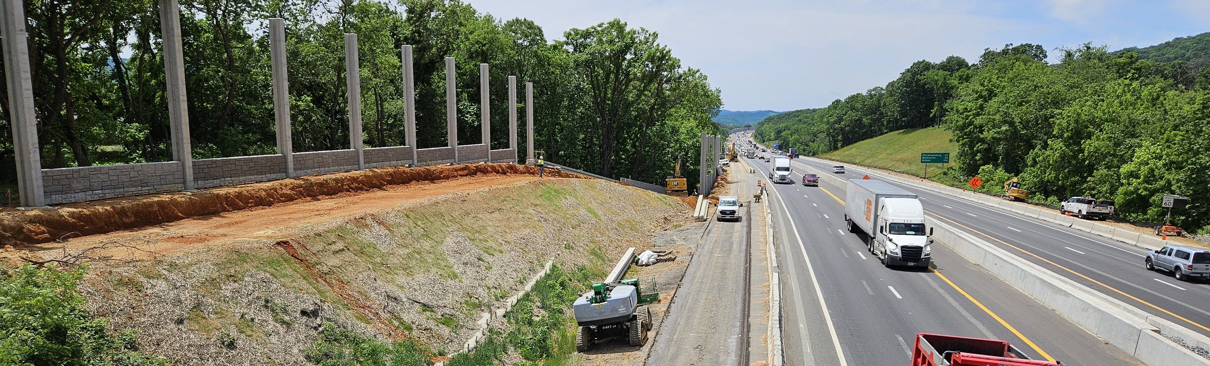 Construction work taking place along a four-lane highway with a mix of car and truck traffic. Work is taking place in the center median and on a sound-barrier wall to the left.
