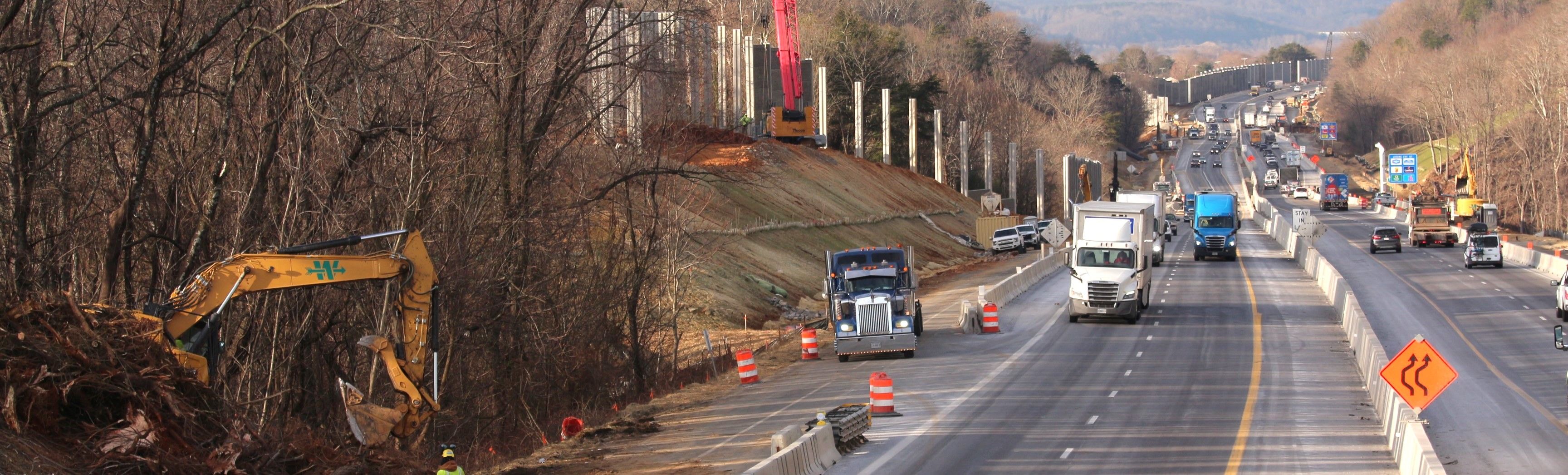 Interstate traffic moving through a construction zone. Concrete barriers are in place along travel lanes. Crews are excavating slopes and constructing noise barriers.