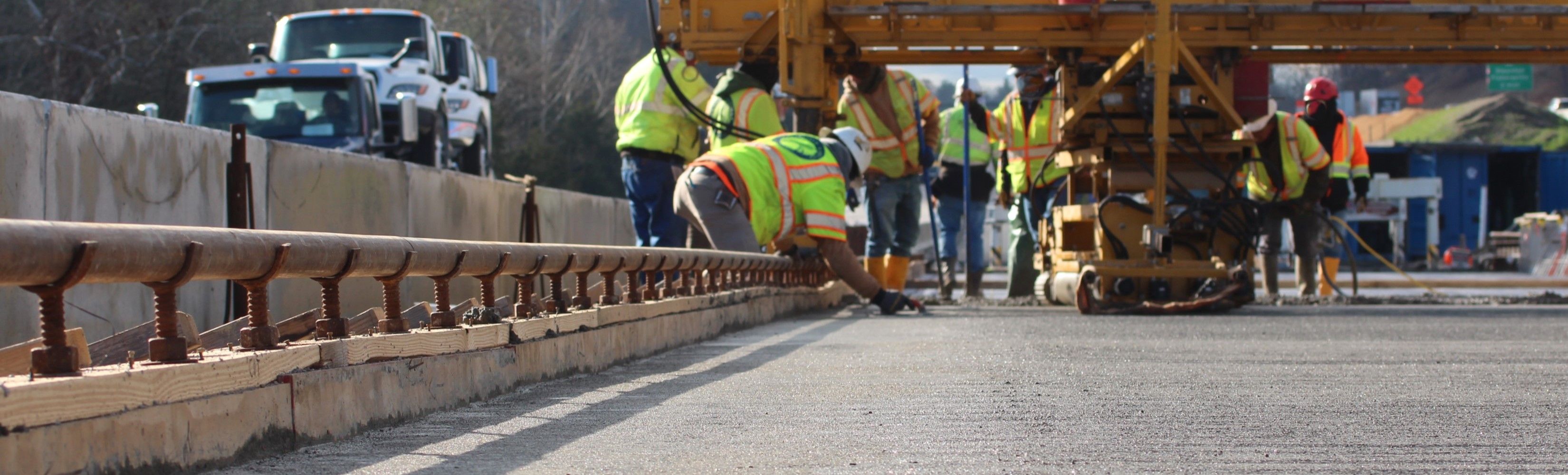Low-angle photo of construction crews pouring concrete for a new bridge deck. Highway traffic is visible to the left.